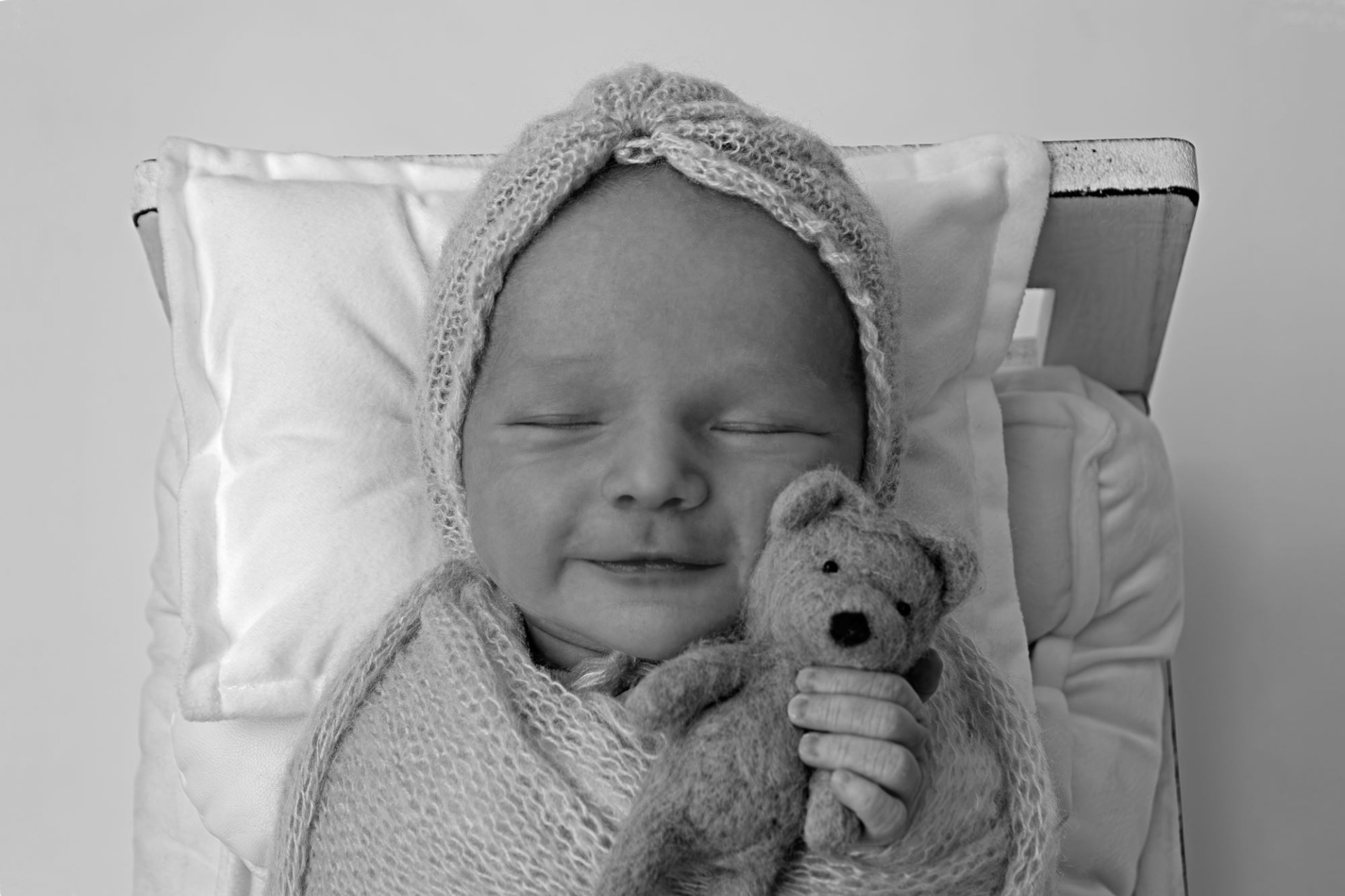 Černobílá fotografie z novorozeneckého, newborn focení. Několika denní miminko drží v ruce malého plyšového plyšáka. Má na sobě modré oblečení a bílé pozadí. Domácí fotoateliér Mladá Boleslav.