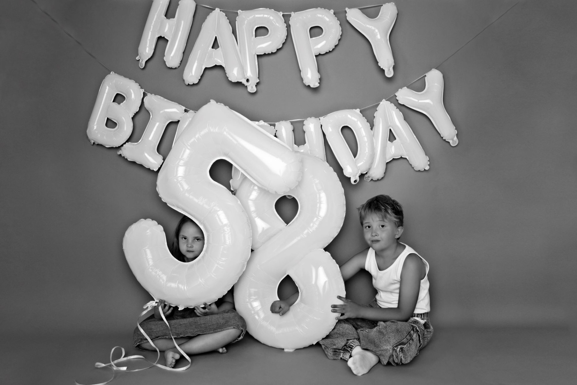 Černobílá fotografie z narozeninového focení. Děti mají balónky s čísly 5 a 8. Za nimi je hnědé pozadí a nápis happy birthday. Domácí fotoateliér Mladá Boleslav.